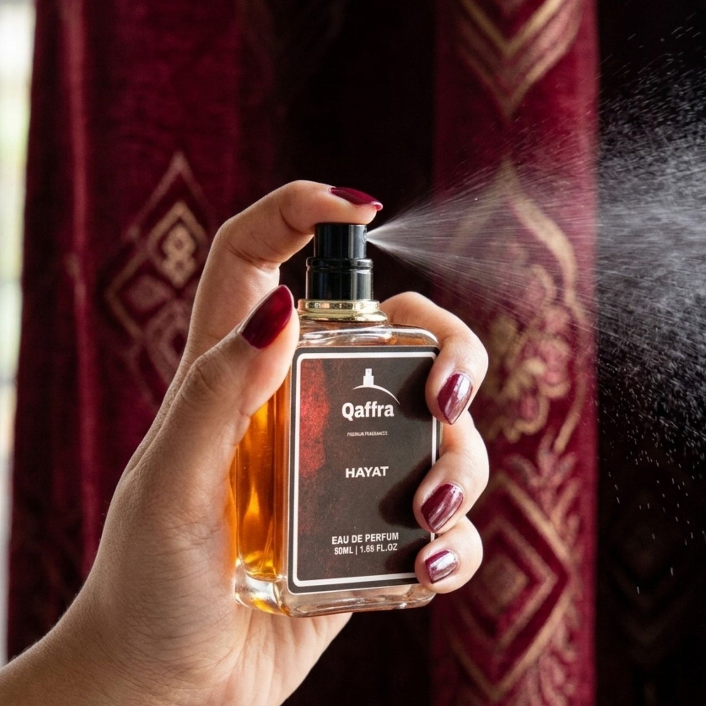 A hand with dark red nail polish holds a bottle of Qaffra Hayat Eau de Parfum, spraying a fine mist. The background features a deep red patterned curtain.