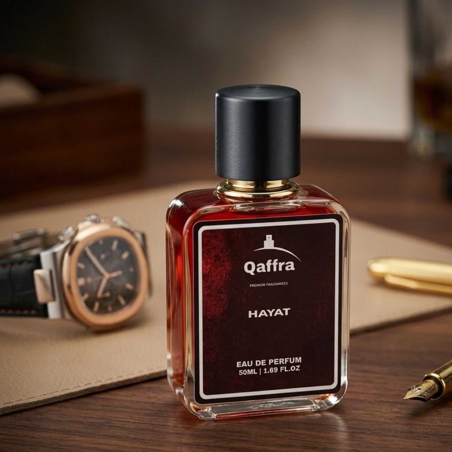 A bottle of Qaffra Hayat Eau de Parfum with a black cap and a dark red-brown liquid inside, displayed on a wooden desk with a watch, leather pad, and gold pen in the background.