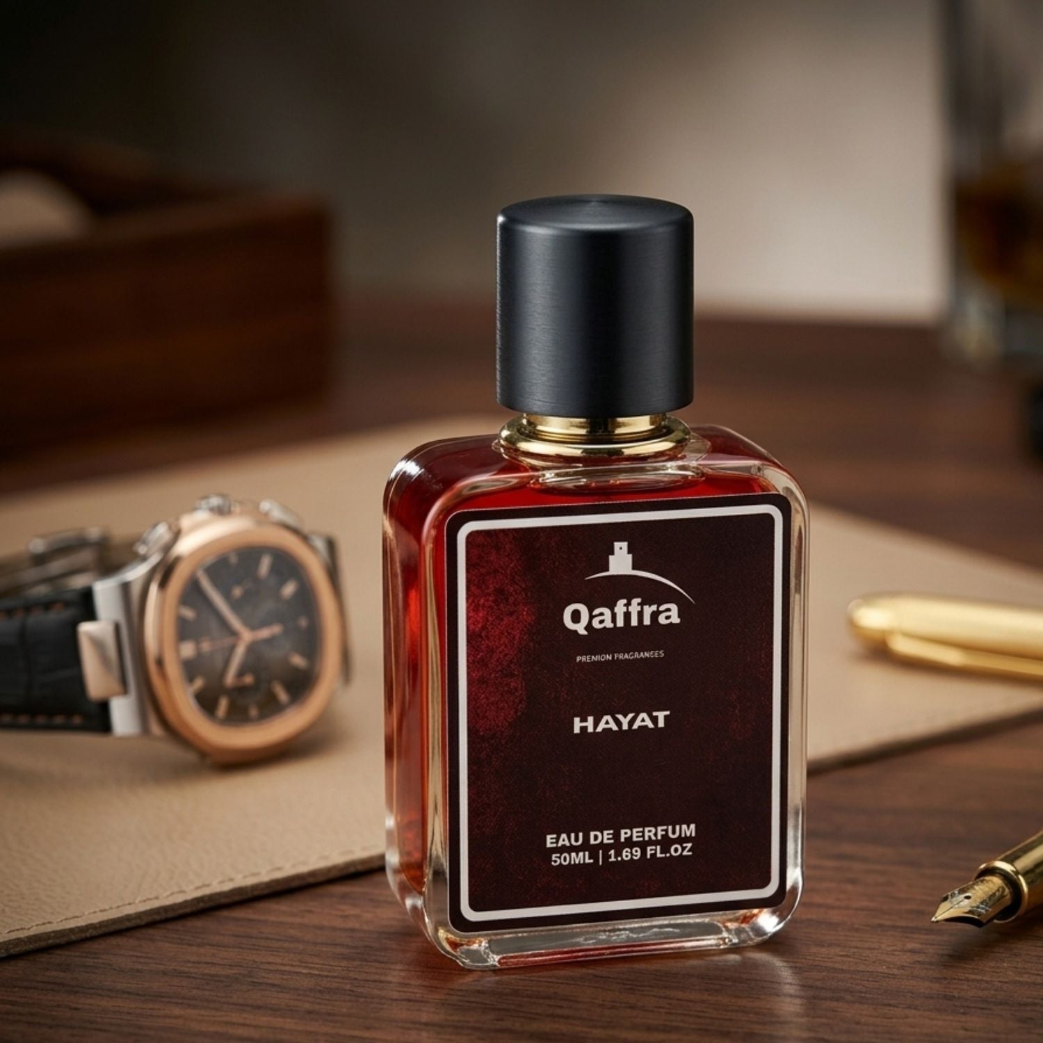 A bottle of Qaffra Hayat Eau de Parfum with a black cap and a dark red-brown liquid inside, displayed on a wooden desk with a watch, leather pad, and gold pen in the background.