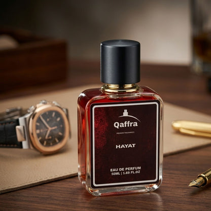 A bottle of Qaffra Hayat Eau de Parfum with a black cap and a dark red-brown liquid inside, displayed on a wooden desk with a watch, leather pad, and gold pen in the background.