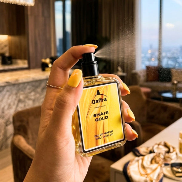 A hand with yellow nail polish holds a bottle of Qaffra Shahi Gold Eau de Parfum, spraying a fine mist in an indoor setting with a blurred city view in the background.