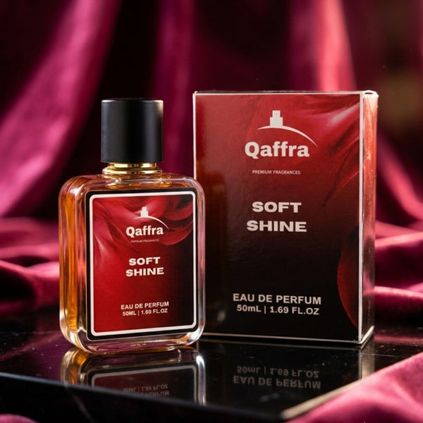 A bottle and box of Qaffra Soft Shine Eau de Parfum displayed together on a reflective black surface with draped magenta fabric in the background.
