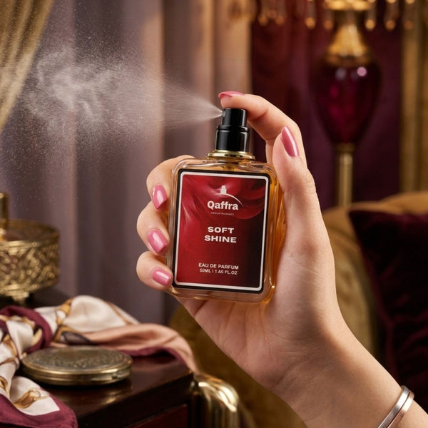 A hand with pink manicured nails holds a bottle of Qaffra Soft Shine Eau de Parfum, with the spray misting out. The background is a luxurious, warm-toned room with velvet cushions and gold accents.