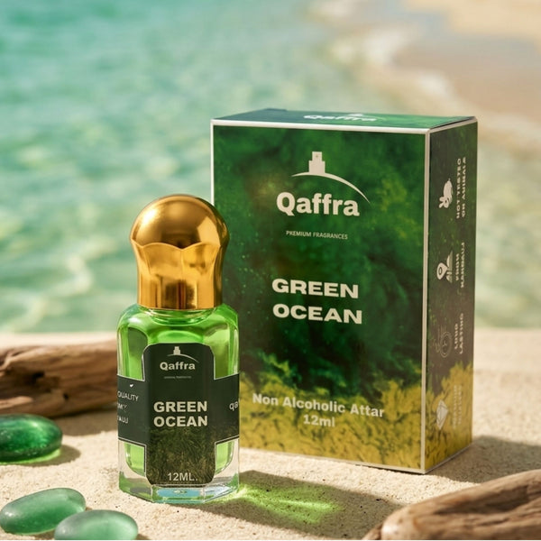 A bottle of Qaffra Green Ocean non-alcoholic attar perfume with a gold cap and green liquid, displayed next to its green and gold packaging box on a sandy beach with turquoise water in the background.